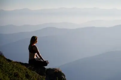 Woman meditating in a seated position on a mountain ledge overlooking misty blue mountain ranges at sunrise.
