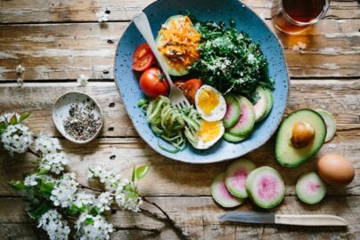 A rustic wooden table set with a colorful healthy bowl containing boiled eggs, spiralized zucchini, cherry tomatoes, avocado, leafy greens, and watermelon radish slices, accompanied by a beverage and spring blossoms.
