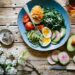 A rustic wooden table set with a colorful healthy bowl containing boiled eggs, spiralized zucchini, cherry tomatoes, avocado, leafy greens, and watermelon radish slices, accompanied by a beverage and spring blossoms.
