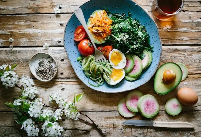A rustic wooden table set with a colorful healthy bowl containing boiled eggs, spiralized zucchini, cherry tomatoes, avocado, leafy greens, and watermelon radish slices, accompanied by a beverage and spring blossoms.