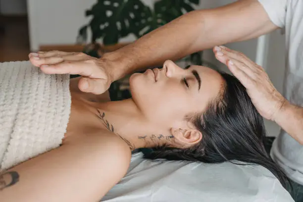 A woman receiving Reiki healing therapy, lying peacefully with eyes closed as a practitioner's hands hover over her head and chest.