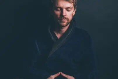 Man sitting cross-legged in meditation with eyes closed, wearing a dark robe, in a calm and focused posture.