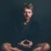 Man sitting cross-legged in meditation with eyes closed, wearing a dark robe, in a calm and focused posture.