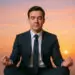 A man in a business suit meditating in a lotus position at sunset, eyes closed, with a calm expression.
