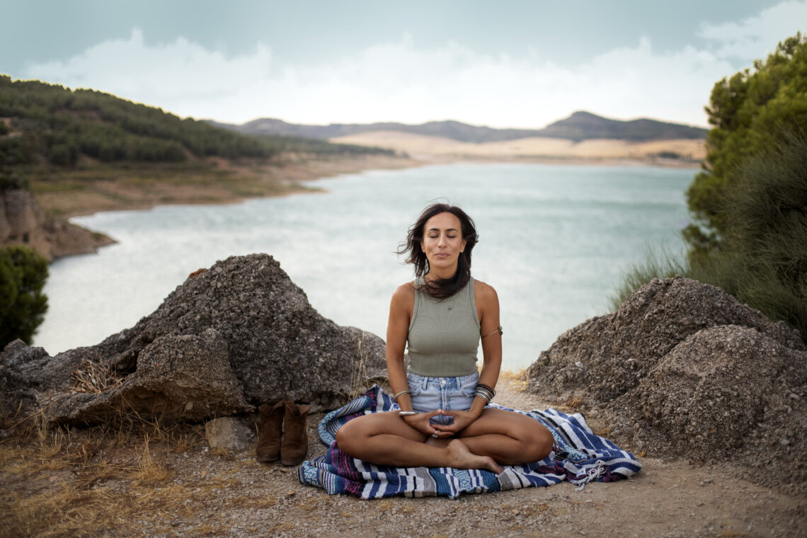 A woman in white meditating in lotus pose on a mountaintop at sunrise, surrounded by serene hills.