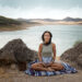 A woman in white meditating in lotus pose on a mountaintop at sunrise, surrounded by serene hills.