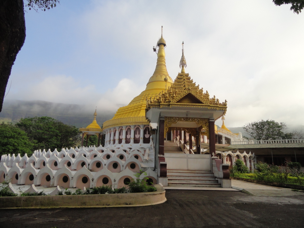 Golden dome of the Global Vipassana Pagoda surrounded by arched structures and trees under a blue sky.