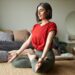 Stylish middle-aged woman practicing meditation in lotus pose at home with eyes closed, wearing a red top and sitting peacefully in a modern bedroom.