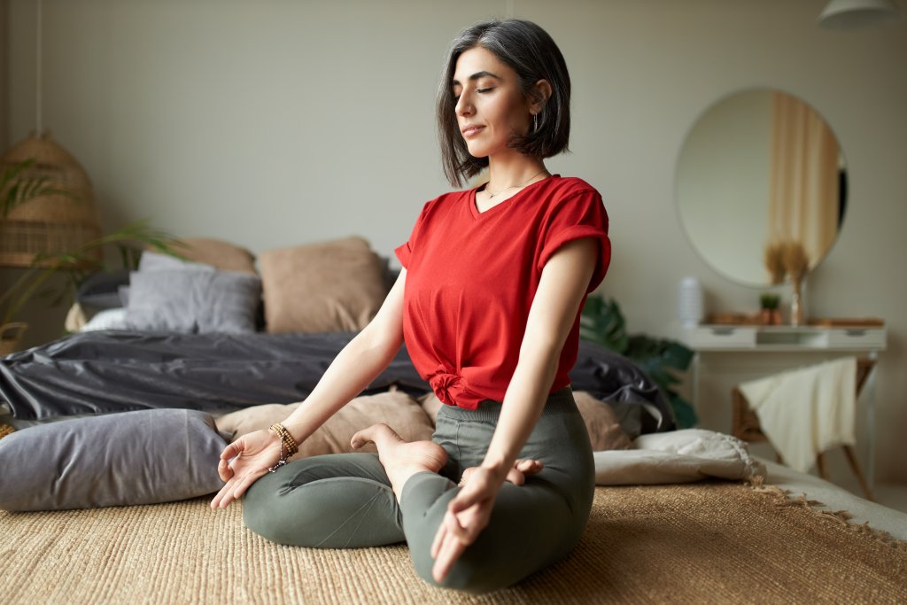 Stylish middle-aged woman practicing meditation in lotus pose at home with eyes closed, wearing a red top and sitting peacefully in a modern bedroom.