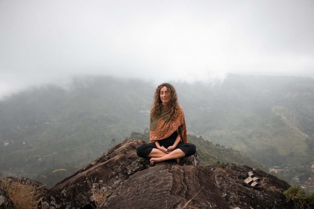 Woman meditating cross-legged on a mountain top with misty hills in the background.