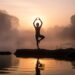 Person practicing yoga on a lakeside at sunrise, surrounded by mist and trees.
