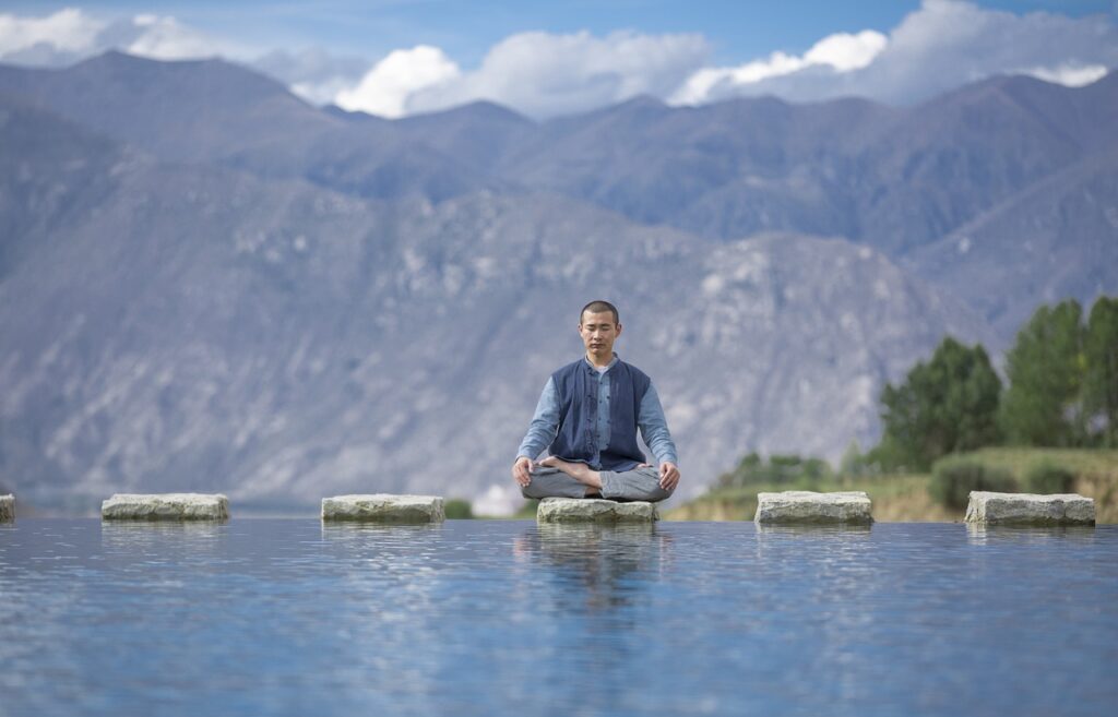 A man meditating in lotus position on a stone slab above water with serene mountains in the background.