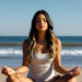 A young woman meditating on the beach in a cross-legged pose, with eyes closed and a calm expression, facing the ocean waves under clear blue skies — symbolizing serenity, mindfulness, and inner peace.