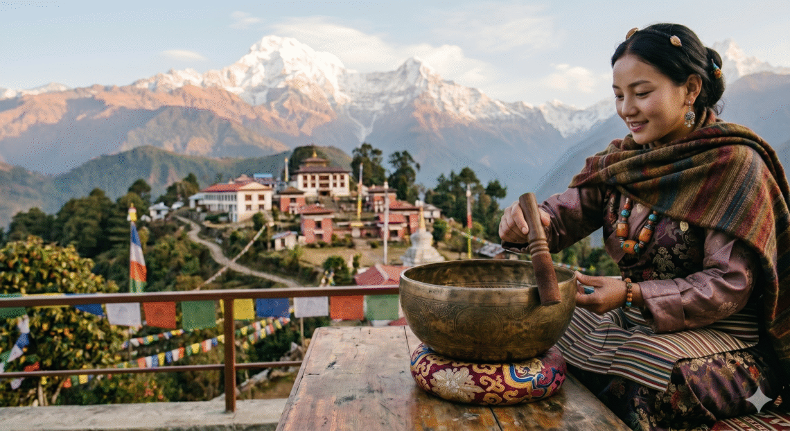 Woman playing a Tibetan singing bowl with a mallet on a terrace overlooking mountains and a monastery in Sikkim.