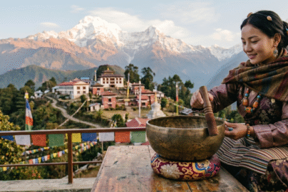 Woman playing a Tibetan singing bowl with a mallet on a terrace overlooking mountains and a monastery in Sikkim.